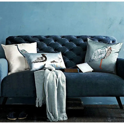 Light blue heron embroidered paradise silk throw pillow cover on dark blue tufted velvet sofa with decorative bird-patterned pillows