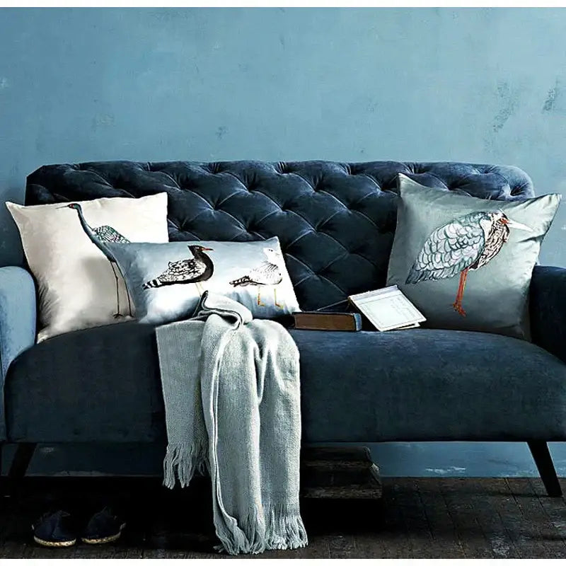 Light blue heron embroidered paradise silk throw pillow cover on dark blue tufted velvet sofa with decorative bird-patterned pillows