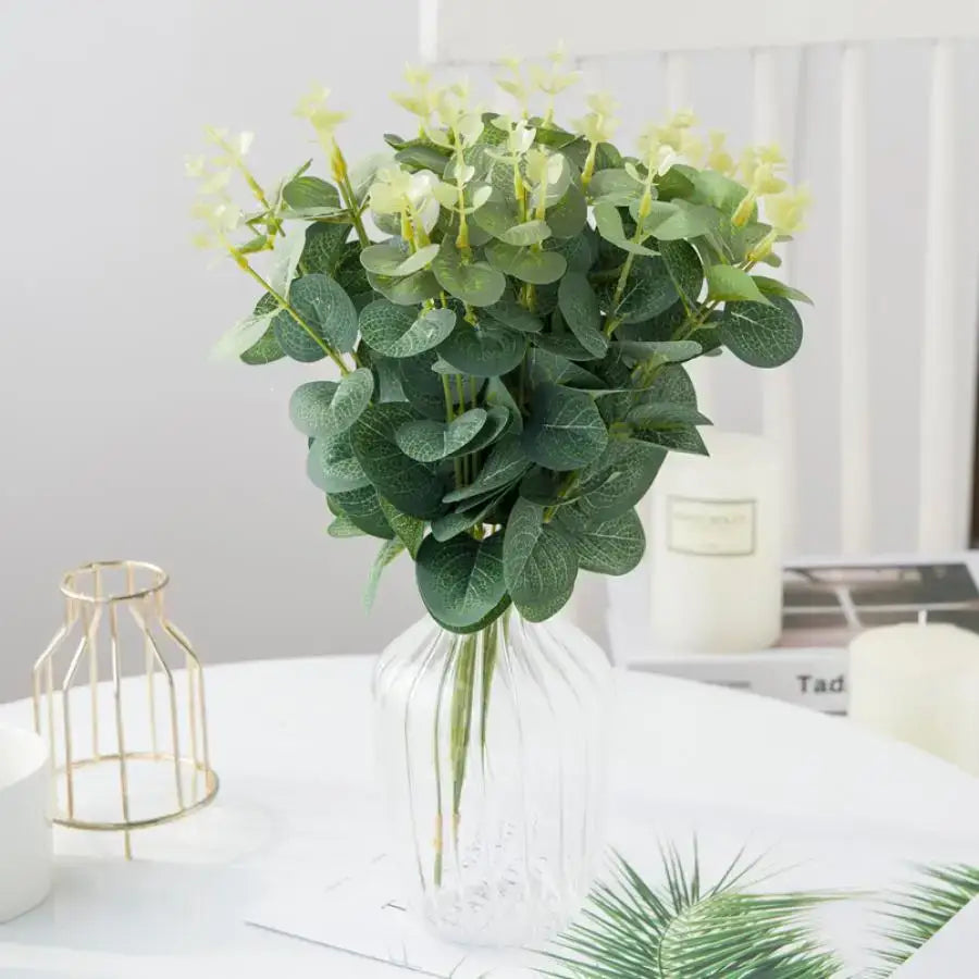 A lush artificial eucalyptus leaves garland with deep green leaves and pale yellow flowers in a clear glass vase for decorbites eucalyptus leaves wedding decor