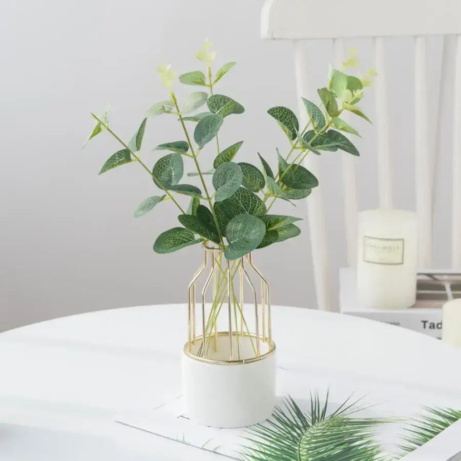 A modern white ceramic vase with gold wire cage design holding decorbites eucalyptus leaves artificial plants for wedding decor