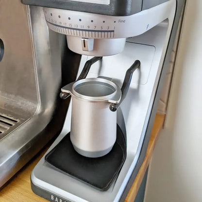 Silver metal milk frothing pitcher with black handles and textured grip base for precise espresso dosing cup use