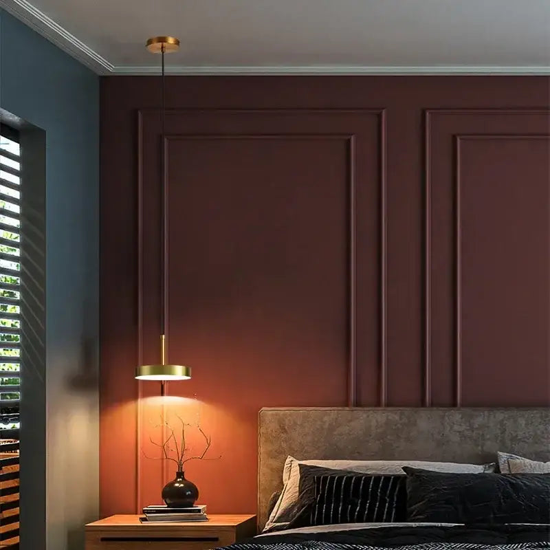 A modern brass pendant light with a frosted white acrylic circular shade hanging above a wooden nightstand