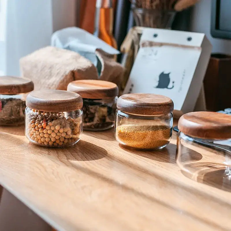 Minimalist clear glass storage jars with smooth rounded acacia wood lids arranged on a light wood surface
