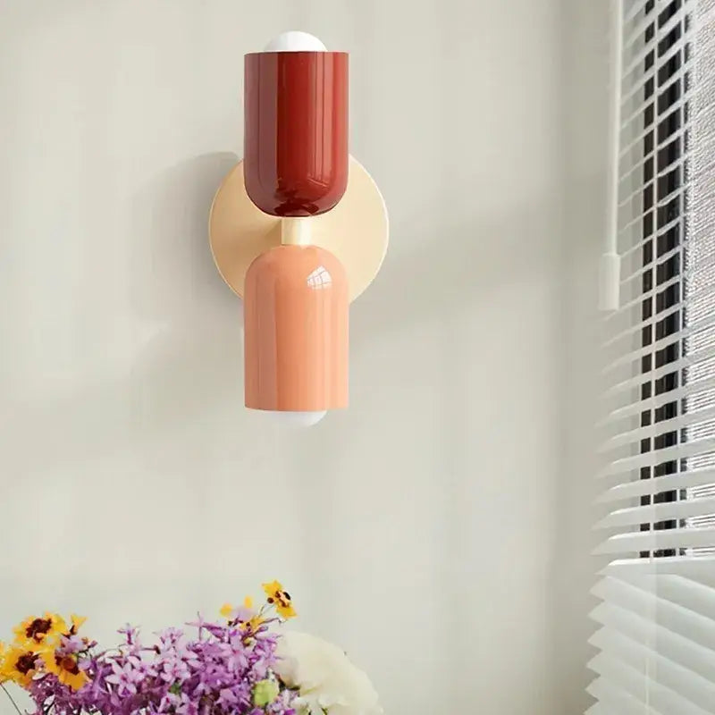 A minimalist cream double head wall lamp with glossy red and orange cylinders on a beige circular backplate