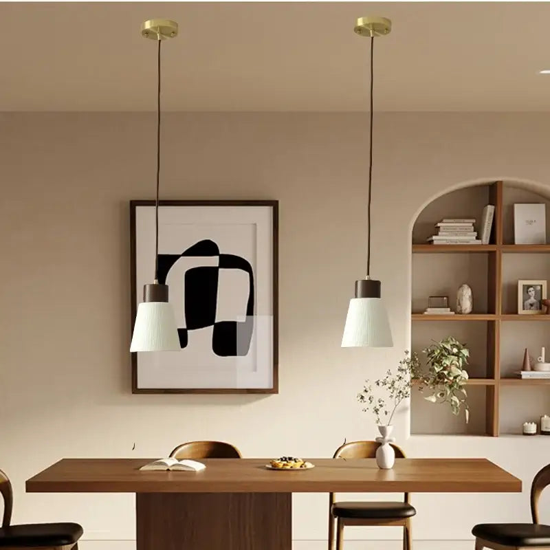 Two minimalist pendant light fixtures with white ribbed glass shades and brass details hang above a wooden table