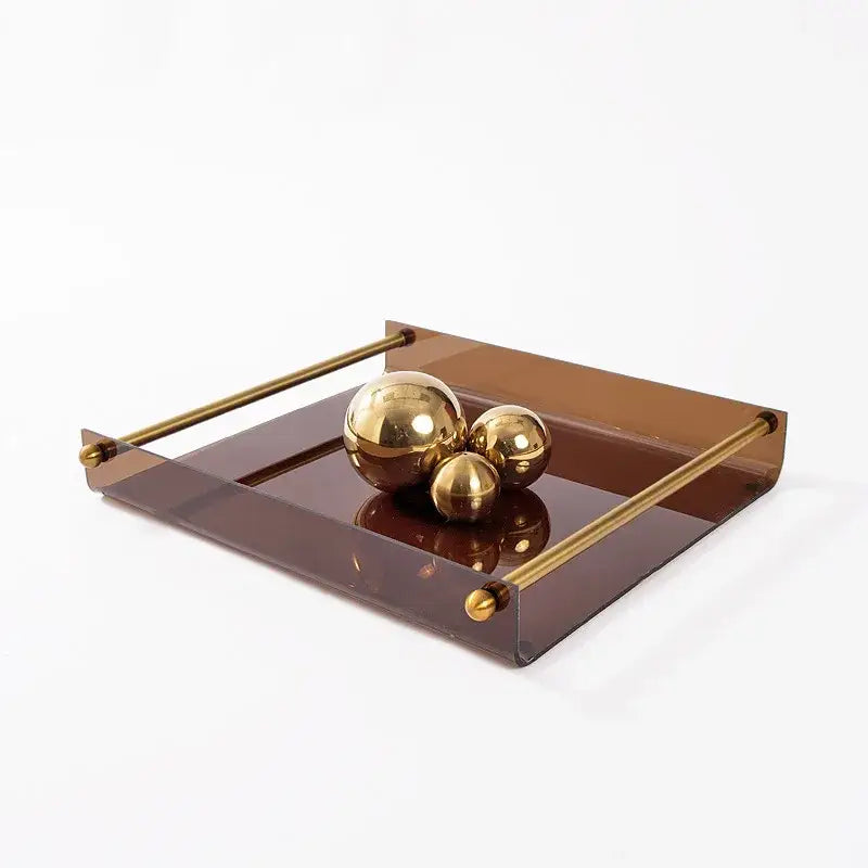 A modern amber acrylic fruit plate tray with polished brass handles holding gold metallic spheres on a reflective surface