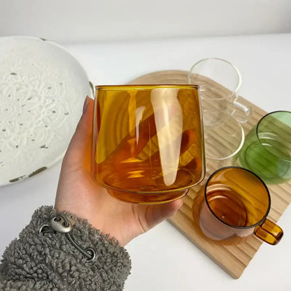 A translucent amber decorbites glass coffee mug with black rim held in hand against colorful mugs background