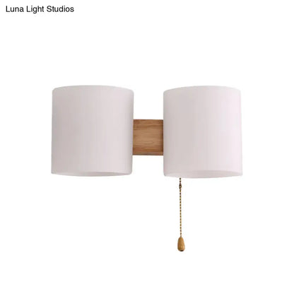 Modern beige fabric and wood wall light bedroom sconce with two cylindrical shades and brass pull chain on natural wood bar
