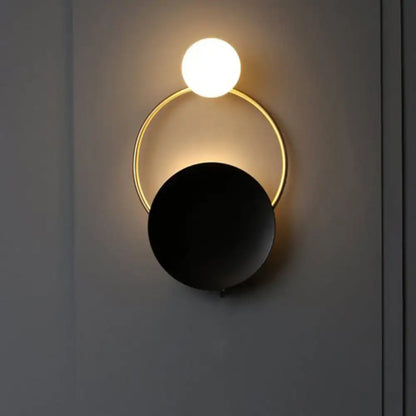 Modern black and gold metal planet wall mount lamp with glowing white spherical bulb and matte black disc bedroom sconce