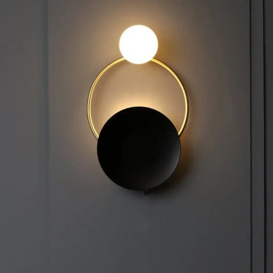 Modern black and gold metal planet wall mount lamp with glowing white spherical bulb and matte black disc bedroom sconce