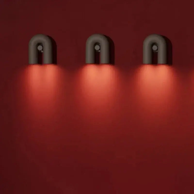 Three sleek matte black and silver tube wall lamps emit a warm red glow in a modern minimalist bedroom sconce design