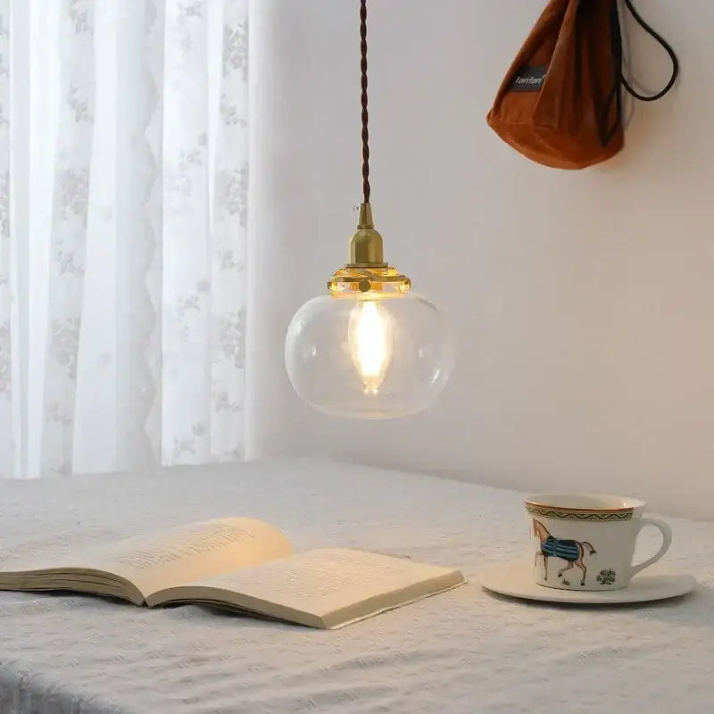 Modern brass and clear glass chandelier pendant light with warm glowing glass and twisted brown cord above an open book