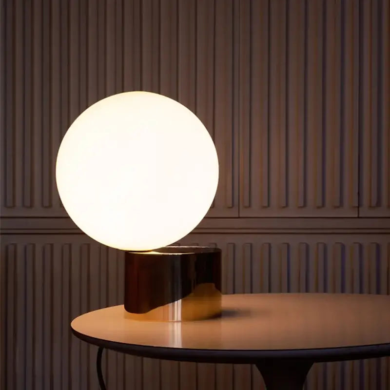 A modern brass and decorbites glass circular table lamp with a glowing white glass circular shade for bedroom use