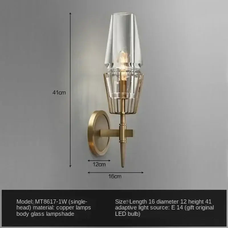 A modern brass and glass bedside table lamp wall sconce with a clear tapered glass shade and metal base