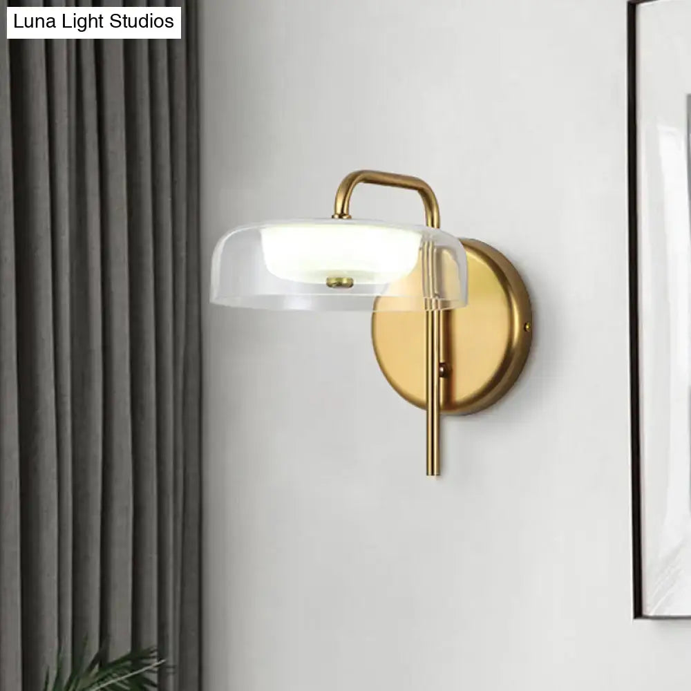 Modern brass and glass bedside wall sconce with gold-toned metal arm and clear dome-shaped glass shade for bedroom lighting
