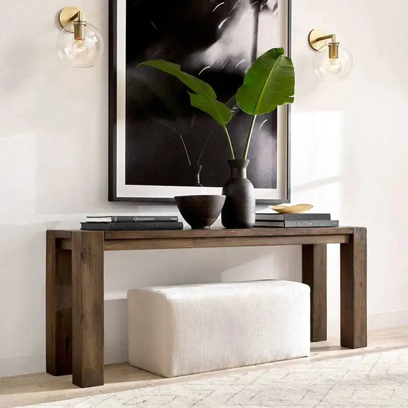 Dark wood console table with thick legs and a black vase with green leaves beneath a modern brass and glass wall sconce bedroom light