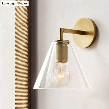 Modern brass and glass wall sconce with a clear glass conical shade and exposed bulb for bedroom lighting