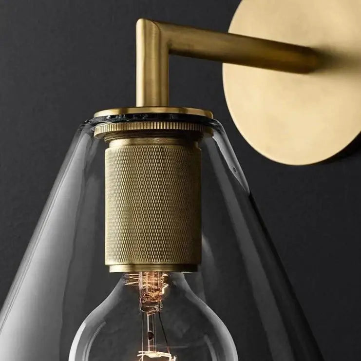 Modern brass and glass wall sconce with textured cylindrical base and clear glass shade housing an exposed filament bulb