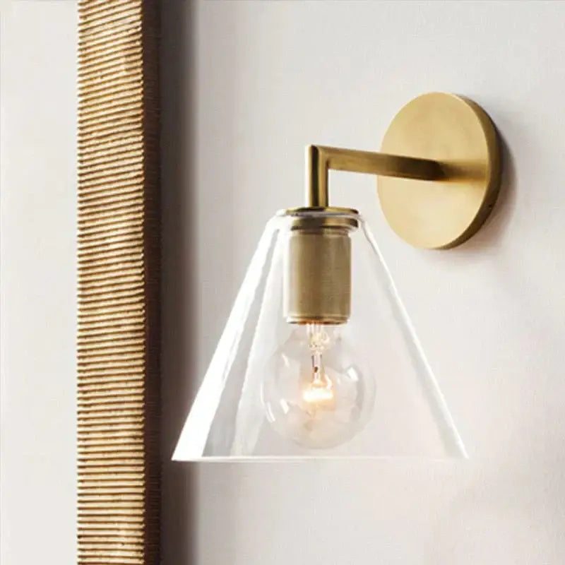A modern brass and glass wall sconce with a clear conical shade and exposed bulb for bedroom lighting