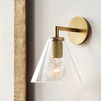 A modern brass and glass wall sconce with a clear conical shade and exposed bulb for bedroom lighting