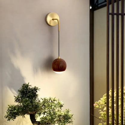 A modern brass and walnut wood wall sconce with a curved arm and spherical wooden shade for ambient bedroom light