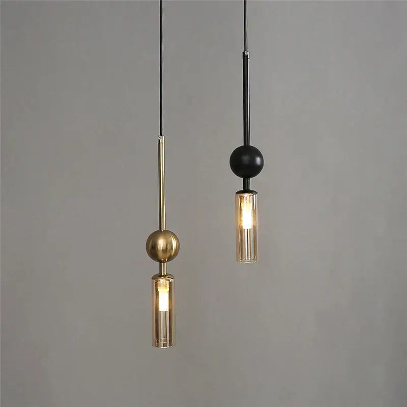 Two modern pendant lights with brass and black finishes feature cylindrical glass shades and spherical accents for dining room use