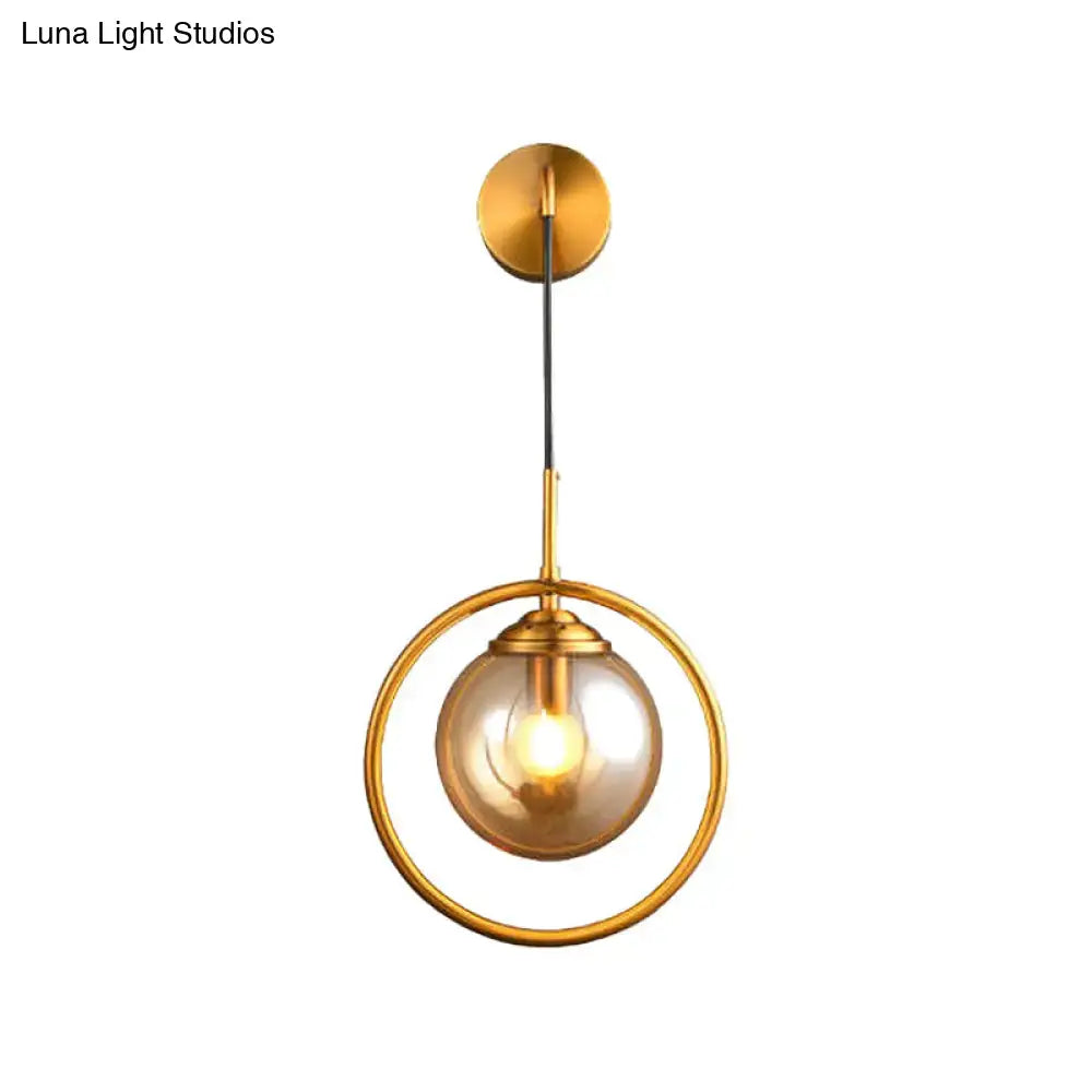Modern brass circular wall mounted lamp with gold-toned metal frame and amber glass globe shade for bedroom lighting