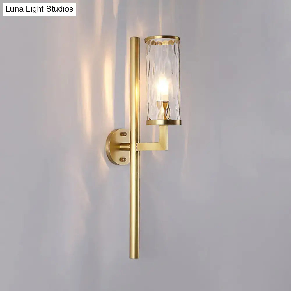 Modern brass cylinder sconce with gold-toned metal arms and clear water glass textured cylinder for bedroom wall lighting
