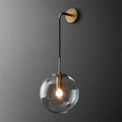A sleek brass-finished modern wall light with a glass ball led globe suspended by a black cord for bedroom decorbites