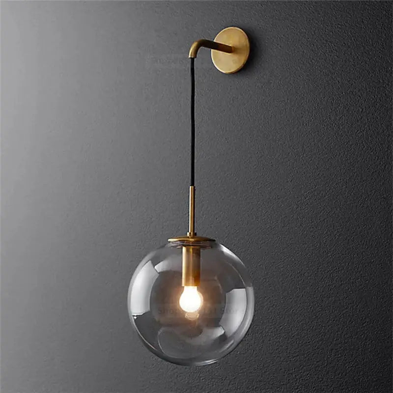 A sleek brass-finished modern wall light with a glass ball led globe suspended by a black cord for bedroom decorbites