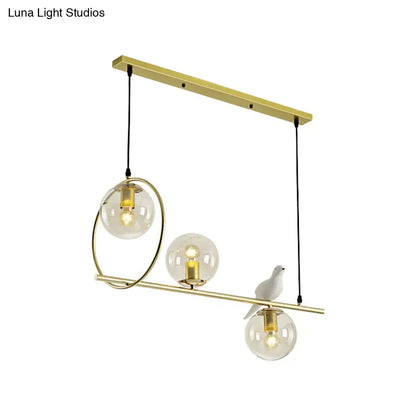 Modern brass global island lamp featuring gold-toned pendant light with three clear glass globes and white bird decor