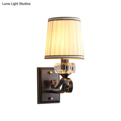 A warm-toned pleated shade on a black brass direct wired electric wall lamp with a dark bronze ornate arm and crystal accent