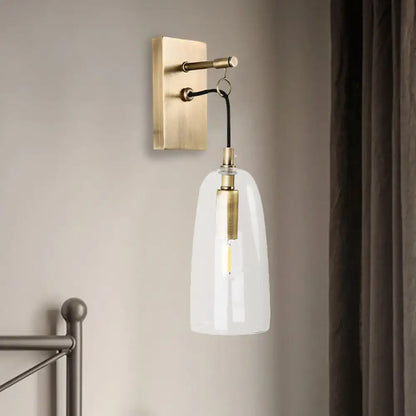 A sleek brass wall mount lamp with a clear glass bell-shaped shade and exposed filament bulb for bedroom use