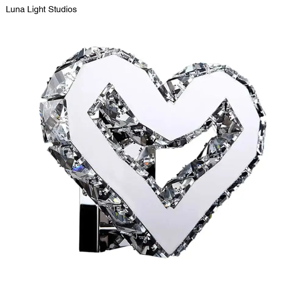 A modern clear crystal heart wall lamp with faceted crystal accents on a chrome base emitting warm white light