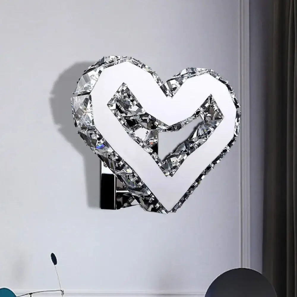 A modern clear crystal heart wall lamp with mirrored facets and chrome base emitting warm white light for bedroom use