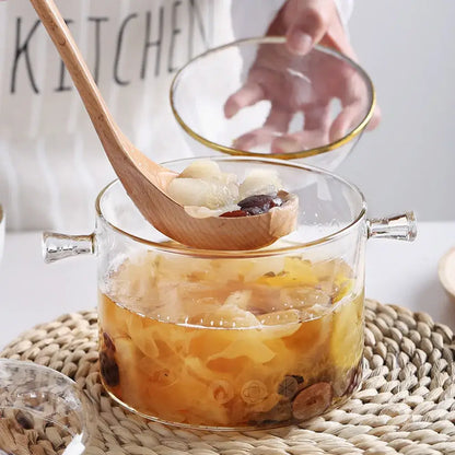 Modern clear glass cooking pot with gold-rimmed handles filled with golden broth and dried fruit ingredients