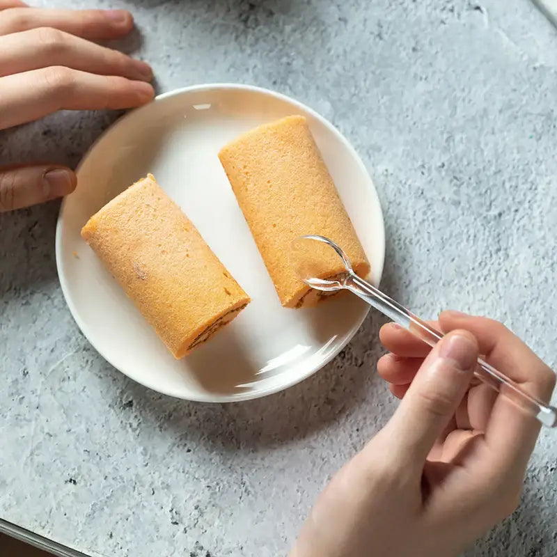 Two golden-brown rolled cakes on a white plate being prodded with a clear glass fork from a modern glass dessert cup set