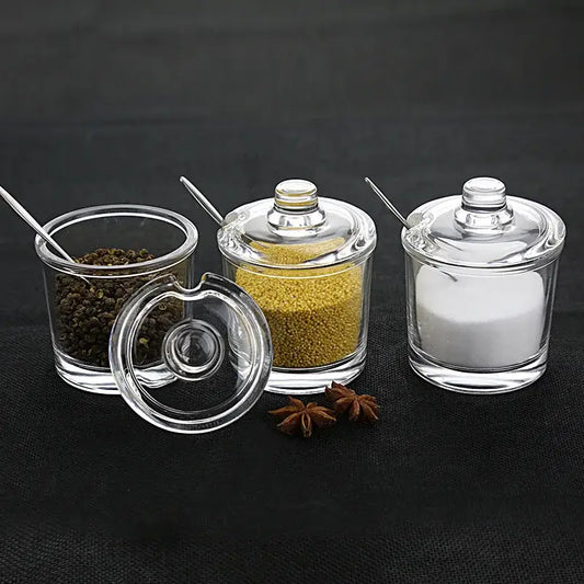 Modern clear glass seasoning jars with lids and metal spoons containing various spices in a spice jar set