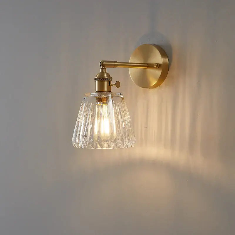 A brass-finished modern bedroom sconce with faceted clear glass shade for stylish wall lights and glass copper wall decor