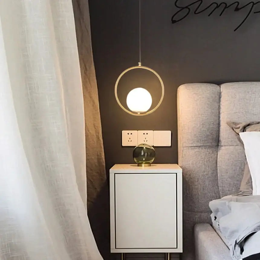 A modern circular glass pendant light with gold metal frame and white glowing bulb for bedroom bedside chandeliers