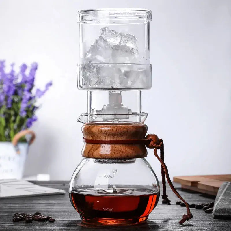Modern glass coffee kettle dripper with wood collar and brown leather strap filled with ice and dark brewed coffee