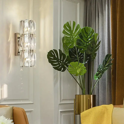 A tall lush green monstera plant in a sleek gold pot beside a modern gold crystal linear wall light dining room sconce