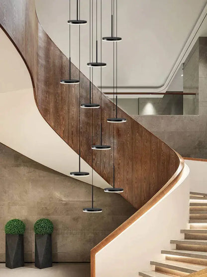 Modern golden black led long chandelier with black circular pendant lights hangs above a curved wooden staircase decorbites golden black
