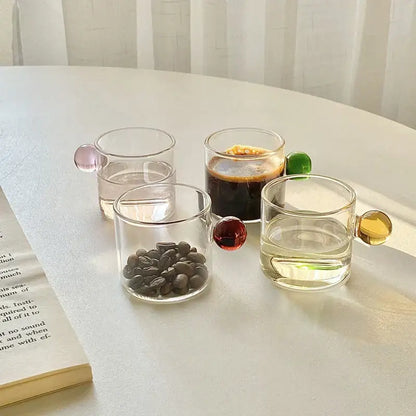 Set of four clear glass espresso cups with colorful pink red and gold spherical handles and marble saucers