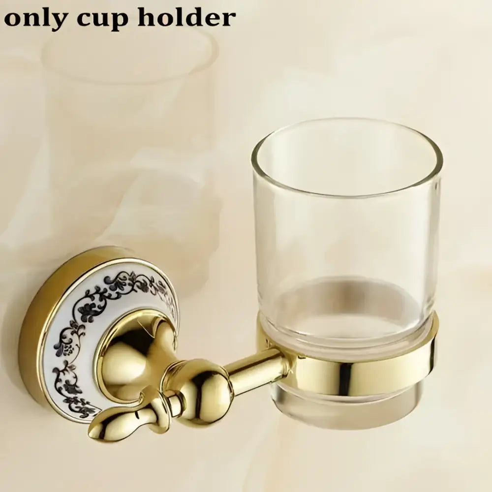 Modern ornate gold and white porcelain bath hardware set featuring a gold-toned cup holder with clear glass tumbler and floral backplate