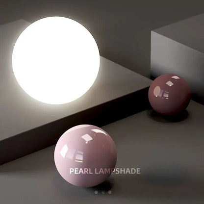A cream nordic led pendant light with a glowing white glass lampshade beside matte pink spheres on a gray surface