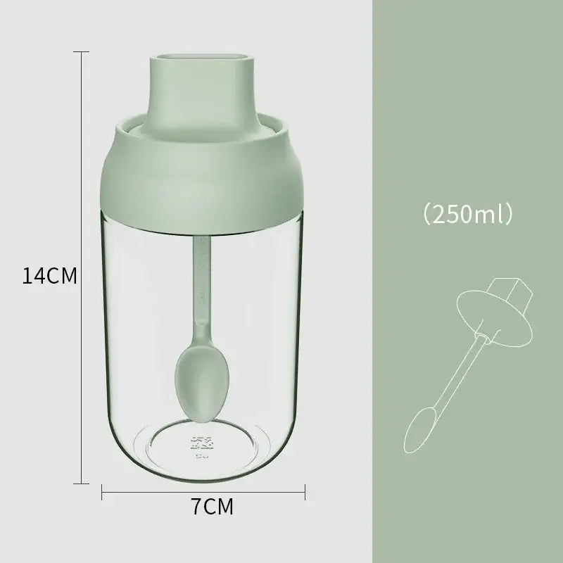 A clear glass seasoning bottle with a matte sage green lid and integrated spoon for convenient kitchen use