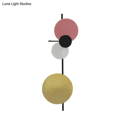 Modern abstract wall sconce featuring three stacked circular discs in matte pink, black, and white, with a brushed gold lower disc mounted on a sleek black rod.