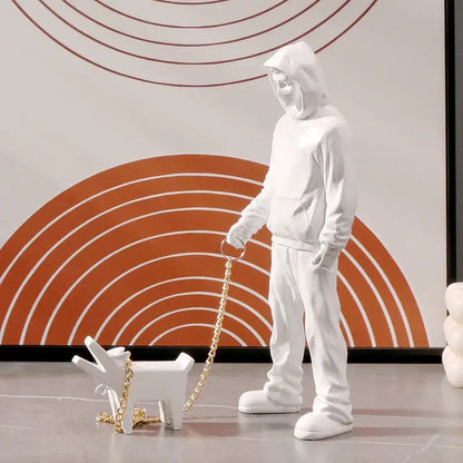 White ceramic decorbites dog sculpture of a person in a hoodie walking a small dog on a gold chain leash for living room decor