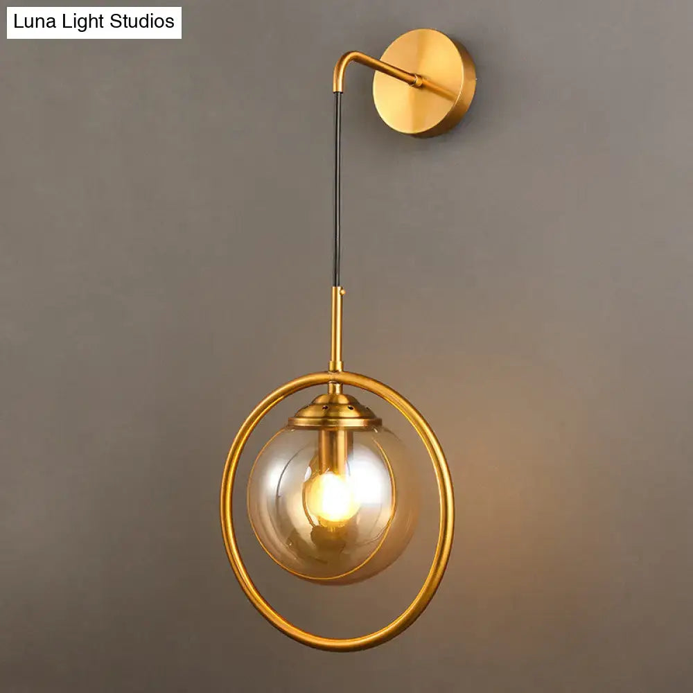 Gold-toned wall sconce with smoke gray clear glass globe encased in ornamental ring suspended by black cord for bedroom lighting